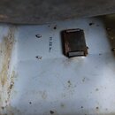 Detail vnitřní strany světlého, pravděpodobně kovového kufru nebo pouzdra. Povrch je znečištěný, s viditelnými skvrnami a oxidací. Uvnitř je umístěn…