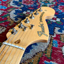 Detail hlavy elektrické kytary z přírodního světlého dřeva. Na hlavě je černý nápis Fender STRATOCASTER a v horní části sériové číslo 1451. Jsou vidě…
