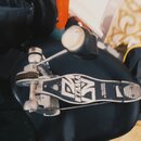 Fotografie zachycuje bicí pedál v černé a stříbrné barvě, který leží na černém povrchu. Na základní desce je vidět bílé logo a nápis TAMA. Povrch ped…