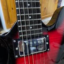 Detail elektrické kytary s červeně-černým přechodem barvy (sunburst). Je vidět hmatník s bílými tečkovými značkami a chromovaný humbucker s viditelný…