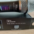 Černý bezdrátový mikrofon s kovovou síťkou položený na černém přijímači. Na těle mikrofonu i na předním panelu přijímače je nápis the t.bone a model…
