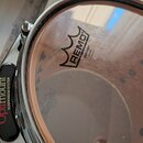 Detailní pohled na průhledný buben s nápisem REMO EMPEROR CLEAR MADE IN USA. Buben je osazen v chromovaném obruči. Na levé straně je vidět černý plas…