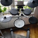 Elektronická bicí souprava značky Roland s černými pady a stříbrnými činely. V horní části je umístěn ovládací modul s nápisem Roland. Na spodním pad…