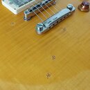 Detailní pohled na tělo elektrické kytary s žlutým flamed maple povrchem. Je vidět kovový most, jedna bílá snímačová lišta a tři hnědé oválné ovládac…