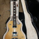 Elektrická kytara typu Les Paul v přírodním dřevě s bílým bindingem, umístěná v otevřeném tvrdém kufru s bílou vnitřní výstuhou. Na hlavě hmatníku je…