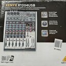 Fotografie znázorňuje krabici od míchacího pultu s nápisem XENYX X1204USB a logem behringer. Na obalu je zobrazen šedý analogový mixér s řadou potenc…