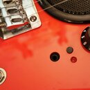 Detail červeného trupu elektrické kytary s kovovým mostem a černým oválným knoflíkem s bílým nápisem VOLUME a čísly 0 až 10. Na povrchu jsou vidět dr…