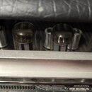 Detailní pohled na čtyři elektronky v zesilovači. Pod nimi je viditelný kovový lem a část čelního panelu s nápisy HANDBUILT IN PETALUMA, CALIFORNIA,…