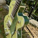 Zelená polodublá elektrická kytara s bílým lemováním korpusu. Na spodní části korpusu je vidět nápis GRETSCH. Nástroj má dva tranzistory, stříbrný ha…