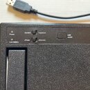 Detailní pohled na horní část černého elektronického zařízení s texturovaným povrchem. Na panelu jsou nápisy Power, Low battery, Joystick, Keyboard,…