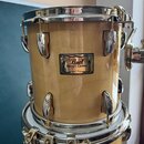 Světle hnědý dřevěný buben s chromovaným obručováním a lugy. Na korpusu je kovový štítek s nápisy Pearl, Masters Custom, MAPLE SHELL a SMALLER textem…