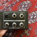 Černý kovový box s konektory a bílými popisky. Horní řada obsahuje výstupy označené jako EMULATED MIC (BAL), LINE (UNBAL) a SPEAKER. Spodní řada pod…