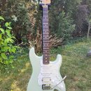 Světle zelená elektrická kytara typu Stratocaster s bílým perleťovým pickguardem a hnedým hmatníkem. Na hlavě je vidět nápis Fender. Nástroj stojí na…