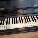 Černé piano s klávesnicí z bílých a černých kláves. Na přední horní části je zlatý nápis AKORD obklopený dvěma čtyřhrotými hvězdami. Pod nápisem je m…
