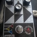 Kytarový efekt pedál s kovovým stříbrným povrchem a černými geometrickými prvky. Na horní části jsou tři černé oválové knoflíky s popisky DELAY, MIX…