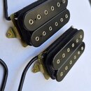 Dvě černé elektrické snímače typu humbucker s kovovými rámečky v mosazném odstínu. Snímače mají plastové kryty s šesti viditelnými póly v řadě. Z kaž…