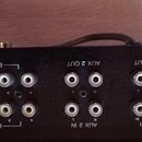 Detailní pohled na zadní panel černého audio zařízení s řadou konektorů. Jsou zde nápisy BUSS OUT, AUX 2 OUT, MON OUT, AUX 1 a AUX 2 IN. Vidět jsou r…