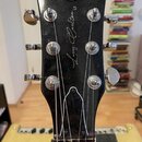 Záhlaví černé elektrické kytary s chromovanými ladičkami. Uprostřed je bílý nápis Larry Carlton L3 a v horní části kulatá nálepka s nápisy PEACE, LOV…