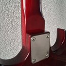 Detail zadní strany červené elektrické kytary s lakovaným povrchem. Je vidět spoj hradla s krkem a kovová krytka upevněná čtyřmi šrouby. Na povrchu n…