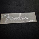 Šedý obdélníkový štítek s bílým nápisem. Je na něm vidět kurzivní logo Fender a pod ním v uvozovkách velká písmena "TELECASTER". Povrch štítku vykazu…