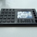 Černý sampler s 16 gumovými pady v mřížce 4x4 a integrovaným displejem. Na horní části je nápis AKAI PROFESSIONAL a na pravém spodním boku modelu MPC…