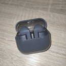 Šedý nabíjecí box s otevřeným víkem, ve kterém jsou umístěny dvě šedé bezdrátové sluchátka. Povrch boxu je matní, s několika drobnými škrábanci a neč…