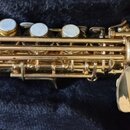 Detailní pohled na část zlatého saxofonu s perleťovými klapkami. Nástroj má lesklý povrch bez viditelných nápisů či značek. Mechanika je komplexní, s…