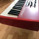 Červený digitální klavír s bílými a černými klávesami. Na červeném povrchu je bílý nápis nord stage 2 a pod ním menší text NORDIC KEYBOARDS. Instrume…