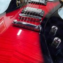 Detail elektrické kytary s červeným plamenovitým topem a černými okraji. Nástroj má dva humbuckery s chromovanými kryty, Tune-o-matic kobylku a stopb…