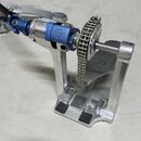 Detailní pohled na kovový mechanismus bubnového pedálu v stříbrné a modré barvě. Na modrém komponentu je vyryto logo ve tvaru stylizovaného kříže v k…