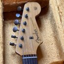 Fotografie zachycuje hlavu elektrické kytary z přírodního světlého dřeva, která leží v plišovém pouzdře. Na hlavě je černý nápis Fender a STRATOCASTE…