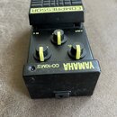Černý efekt pedál s nápisem COMPRESSOR na horní části a YAMAHA CO-10M.II na spodní hraně. Zařízení má tři černé ovládací knoflíky s žlutými značkami…
