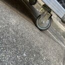 Detailní pohled na spodní část černého transportního kufru s hliníkovými hranami. Na spodní straně jsou vidět dvě gumové kolečka na kovových držácích…
