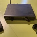 Černý obdélníkový přijímač s displejem a ovládacím knoflíkem. Vedle něj je umístěn černý vysílač s nápisy SENNHEISER a ew G4. V pravé dolní části je…