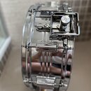Fotografie zachycuje detail vrchu chromovaného malého bubnu. Je vidět kovový lem, napínací šrouby a mechanismus throw-offu. Povrch je vysoce lesklý s…
