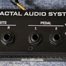 Zadní strana černého kovu s nápisem FRACTAL AUDIO SYSTEMS. Jsou zde vidět vstupy a výstupy s popisky INPUTS (L/Mono, R), OUTPUTS (L/Mono, R) a PEDAL…