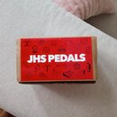 Kartonová krabice s červeným obalem, na kterém je bílý nápis JHS PEDALS. Povrch krabice je doplněn множеstvem drobných bílých ikon a symbolů. Rohy kr…