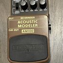 Hnědý efekt pedál s nápisy BEHRINGER, ACOUSTIC MODELER a AM100. Horní část obsahuje tři černé oválné ovladače s popisky MIN a MAX, přepínač MODE (STA…