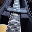 Kytara v černé pouzdře s popruhem, na kterém je bílý nápis Epiphone. Nástroj má světlé tělo, tmavý hmatník s bílými vložkami a kovové pražce. Na hlav…