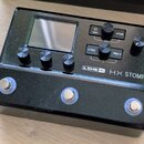 Černý efektový pedál s nápisem LINE 6 HX STOMP. Zařízení má malý displej, pět otočných knoflíků a tři stoupací spínače s modrým podsvícením. Na horní…