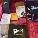 Sada příslušenství značky Gibson na červeném plyšovém podkladu. Obsahuje černý popruh s logem Gibson, černý textilní sáček s logem Gibson a černý man…