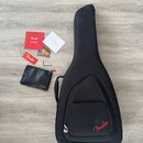 Černý polotvrdý obal na kytaru s červeným logem Fender. Vedle obalu je černý pouzdro s červeným logem Fender, imbusový klíč a několik papírových doku…