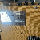 Detail zadní strany zesilovače s žlutým texturovaným potahem. Na povrchu je připevněn kovový štitek s nápisy Blues Junior, FENDER MUSICAL INSTRUMENTS…