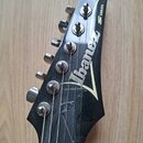 Detail hlavy elektrické kytary v černé barvě s kovovými ladičkami. Na povrchu je bílý nápis Ibanez a v horní části nápis S SERIES. Černý lak vykazuje…