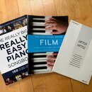 Na fotografii jsou tři knihy položené na dřevěné podlaze. První dvě jsou zpěvníky s nápisem THE REALLY BIG REALLY EASY PIANO SONGBOOK a THE EASY PIAN…