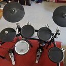 Elektronická bicí souprava s černými pady a činely na černém kovovém rámu. Na modulu je nápis Roland, stejně jako na bílém rámech basového bubnu. Sou…