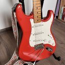 Červená elektrická kytara typu Stratocaster s bílým pickguardem a světle hnědým javorovým hmatníkem. Nástroj má tři snímače, tři ovládací knofíky a c…