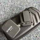Na černém textilním pouzdře leží dva černé plastové moduly bezdrátového systému. Menší modul nese nápisy SYNCO Receiver, zatímco větší modul obsahuje…
