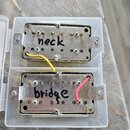 Dvě elektrické kytarové snímače typu humbucker s kovovými kryty v průhledné plastové krabičce. Na horním snímači je ručně napsaný nápis neck a na dol…