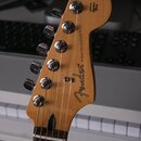 Detail hlavy elektrické kytary z přírodního dřeva. Na povrchu je černý nápis Fender a STRATOCASTER® s doplňkem ORIGINAL Contour Body. Kytara má šest…